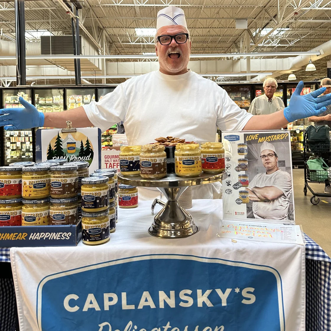 Zane Caplansky in chef's whites behind a Caplansky's demo table stacked with mustard jars
