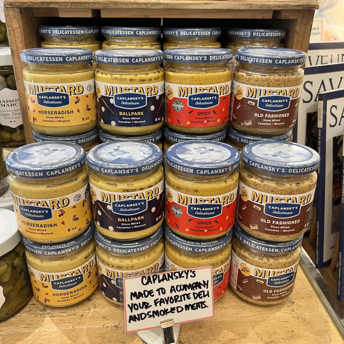 All four Caplansky's mustards stacked in a wooden crate display on a deli counter